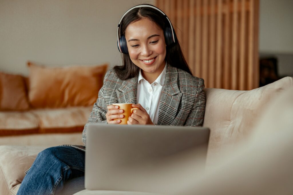 Attractive asian business woman listening music on headphones sitting at laptop with coffee
