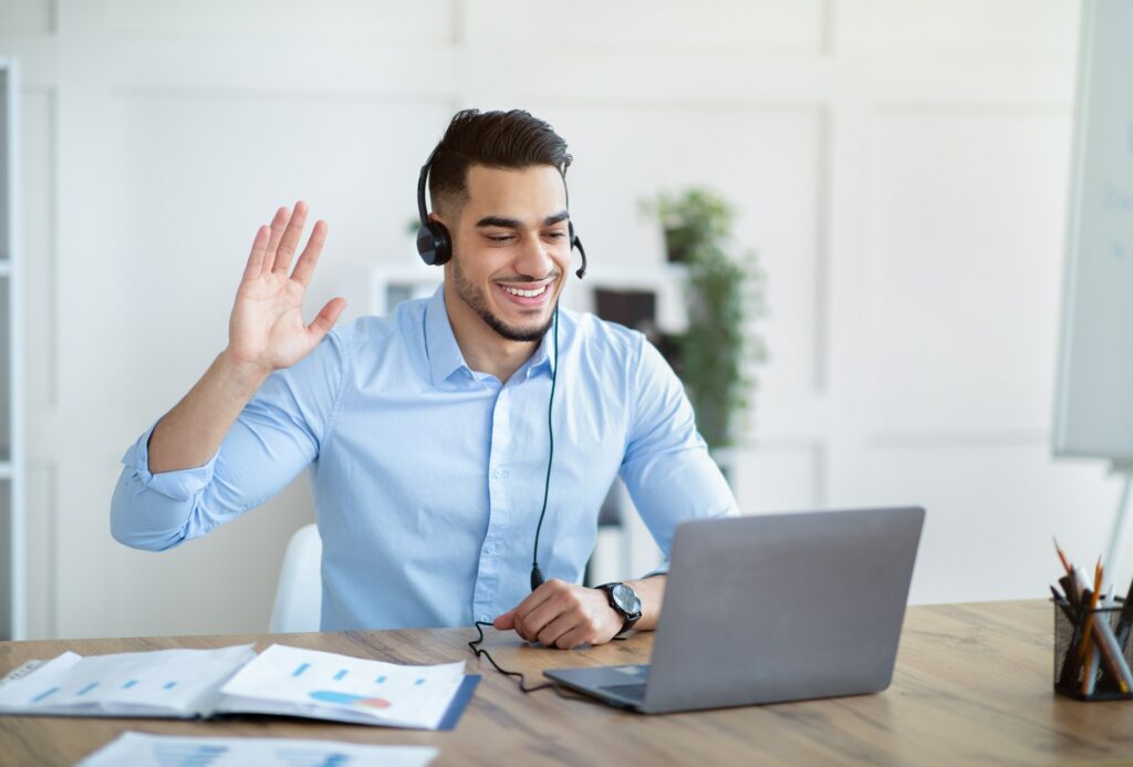 Smiling Arab guy in headset speaking with business partner, having online video call, waving at